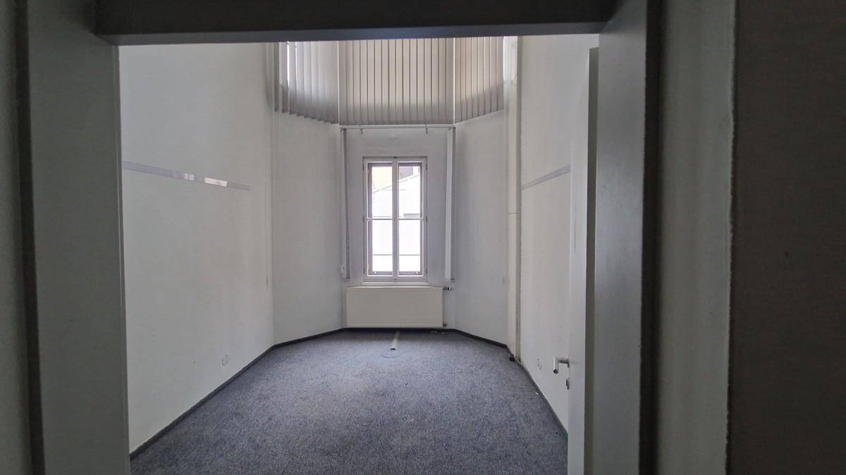 Leerer Büroraum mit Teppichboden und einem Fenster, das durch eine Türöffnung sichtbar ist.