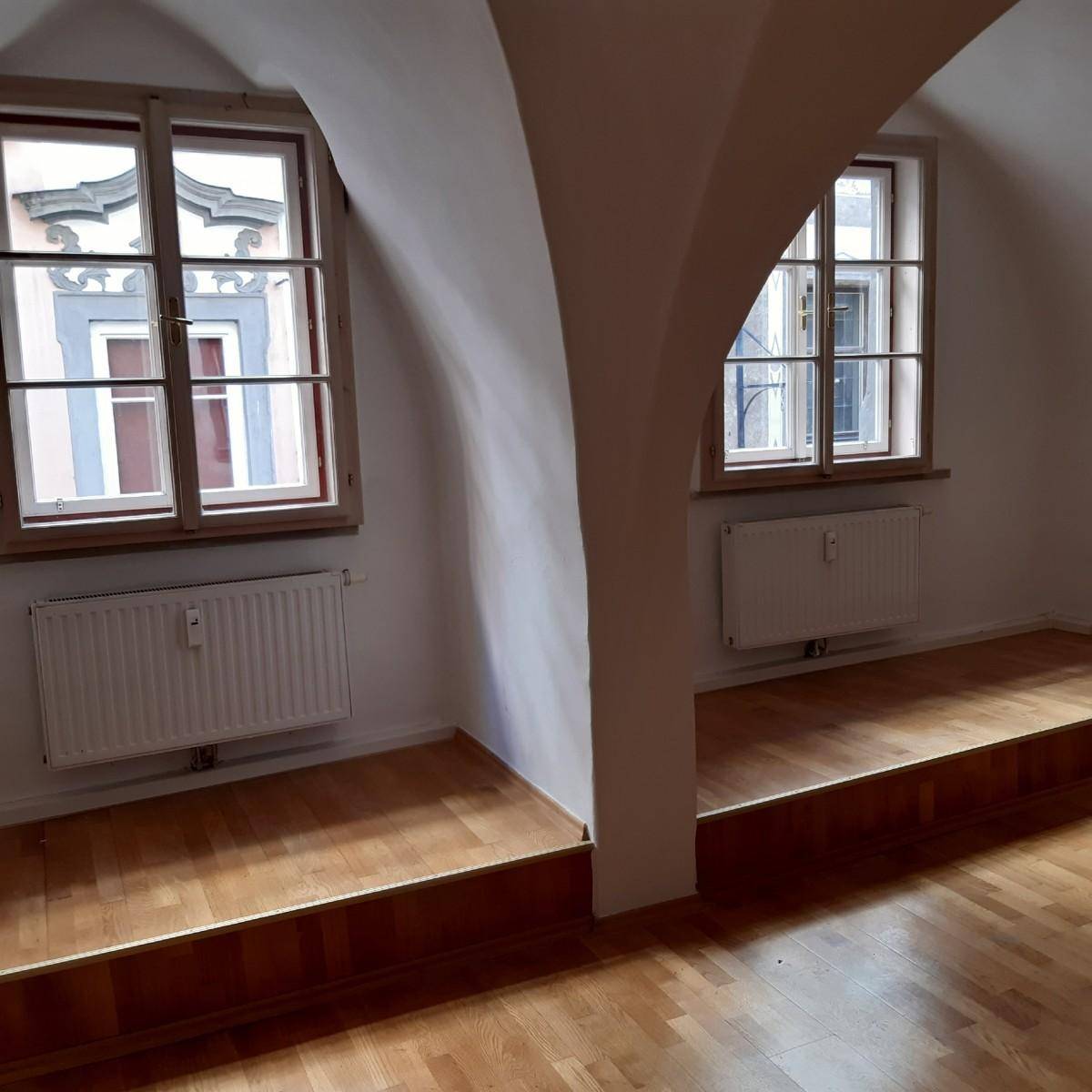 Heller Raum mit Holzboden, Gewölbedecken und zwei Fenstern, geteilt durch einen Bogen.