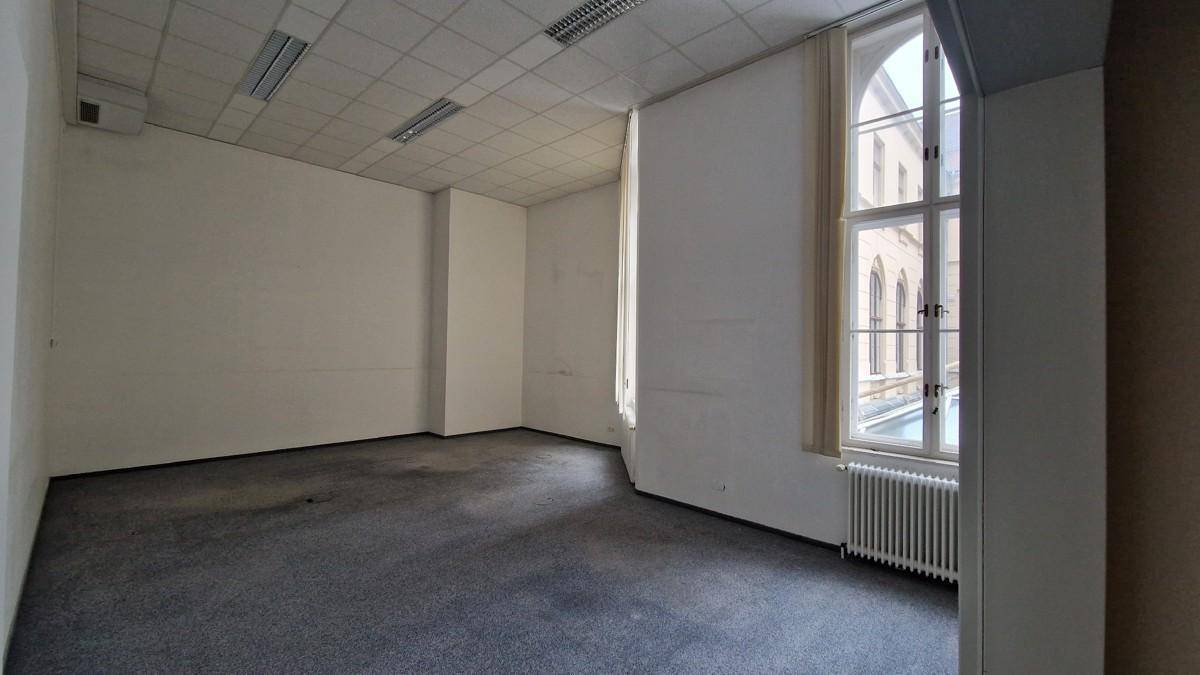 Leerer Büroraum mit Teppichboden, hohen Decken und einem großen, bogenförmigen Fenster, das Tageslicht hereinlässt.