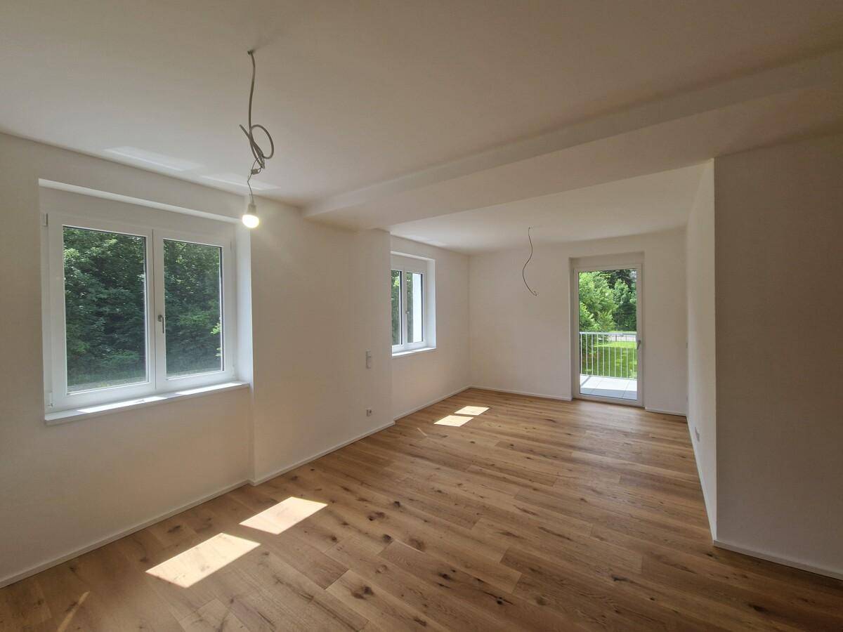 Lichtdurchflutetes Wohnzimmer mit Holzplankenboden, großen Fenstern und Balkonzugang, sehr hell.
