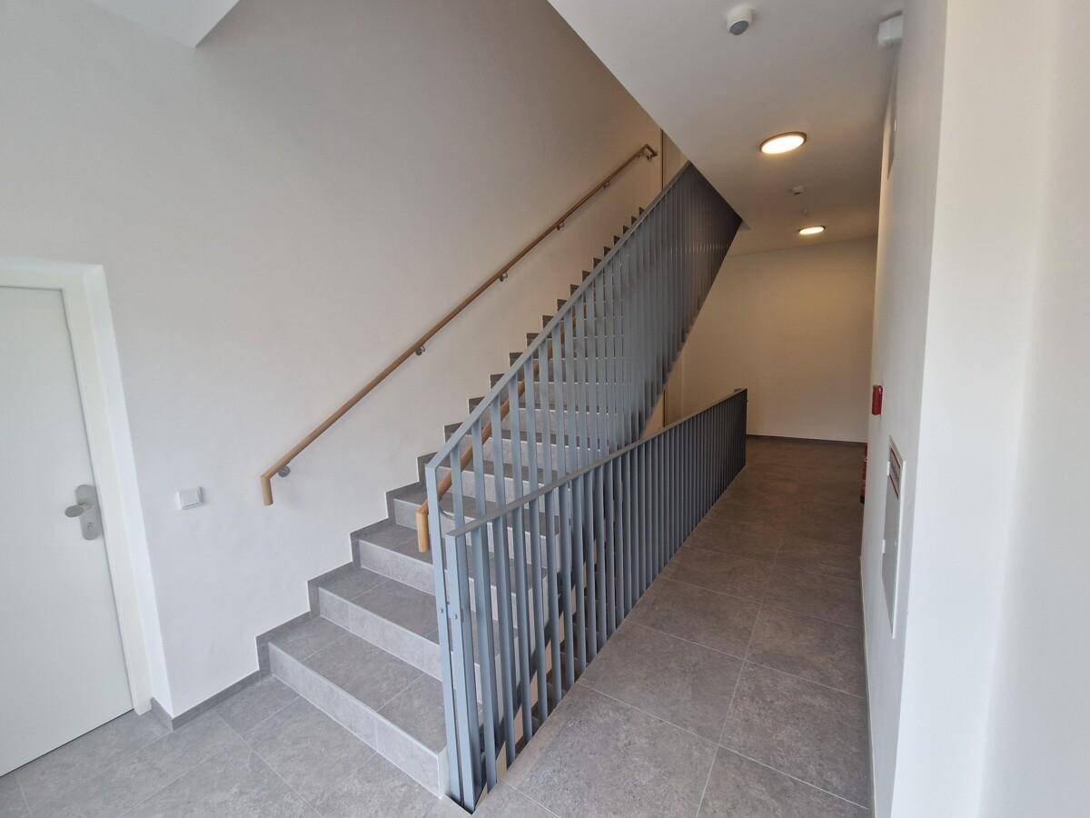 Modernes Treppenhaus mit grauen Fliesen, Metallgeländer und Holzhandlauf in einem Mehrfamilienhaus.