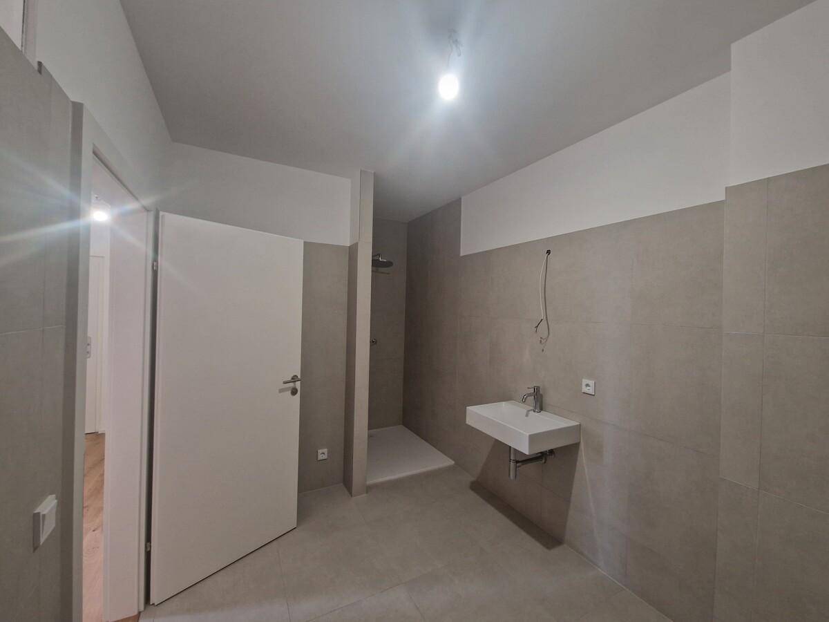 Modernes Badezimmer im Rohbau mit grauen Fliesen, wandhängendem Waschbecken und begehbarer Dusche.