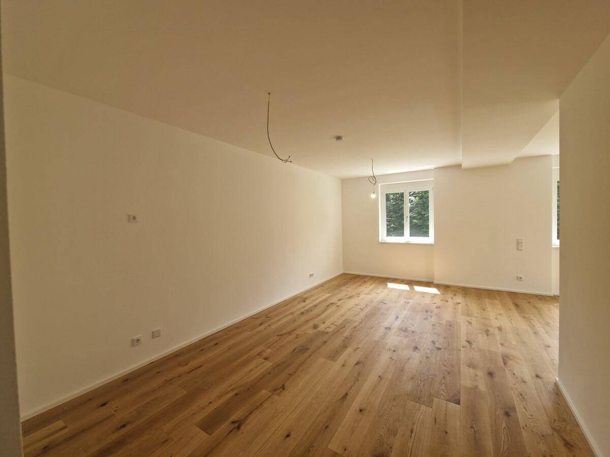 Geräumiges Wohnzimmer mit hellem Holzplankenboden und großen Fenstern, die viel Sonnenlicht hereinlassen.