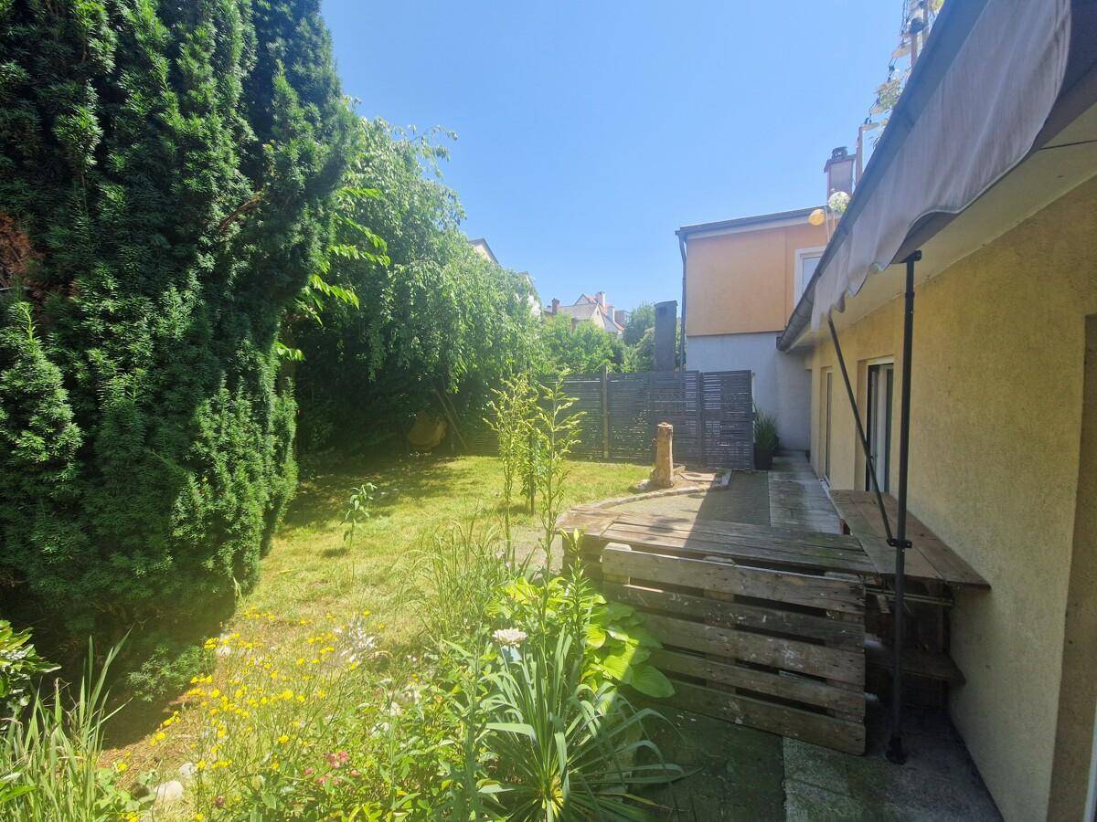 Gartenbereich mit viel Grün, einer Holzterrasse und Blick auf die Rückseite des Gebäudes.