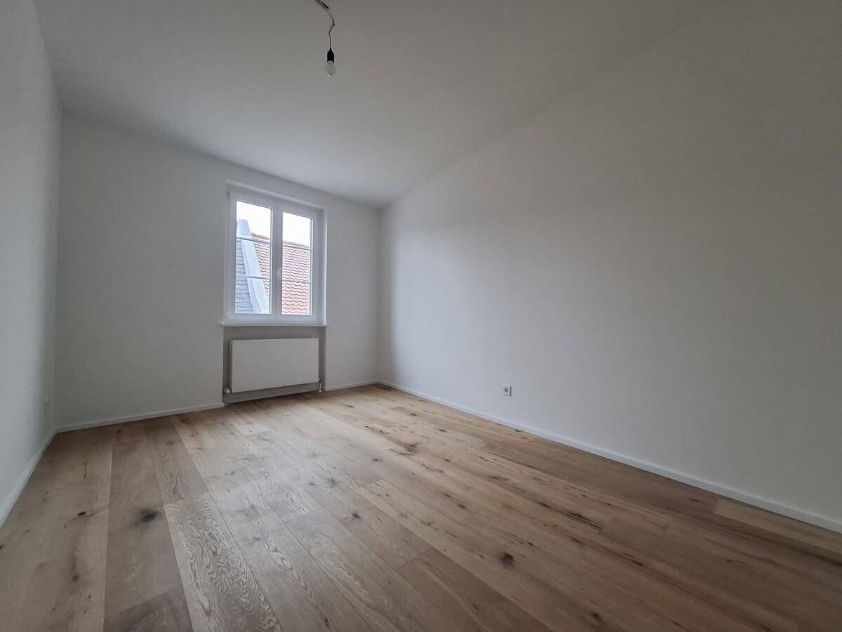 Heller, leerer Raum mit Holzboden und einem Fenster, das Tageslicht hereinlässt.