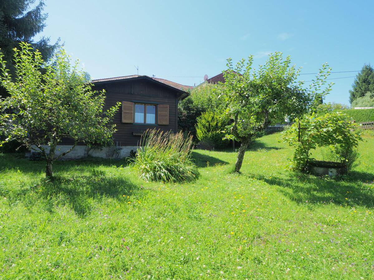 Kleines Holzhaus mit Fensterläden, umgeben von einem gepflegten Rasen und jungen Obstbäumen.