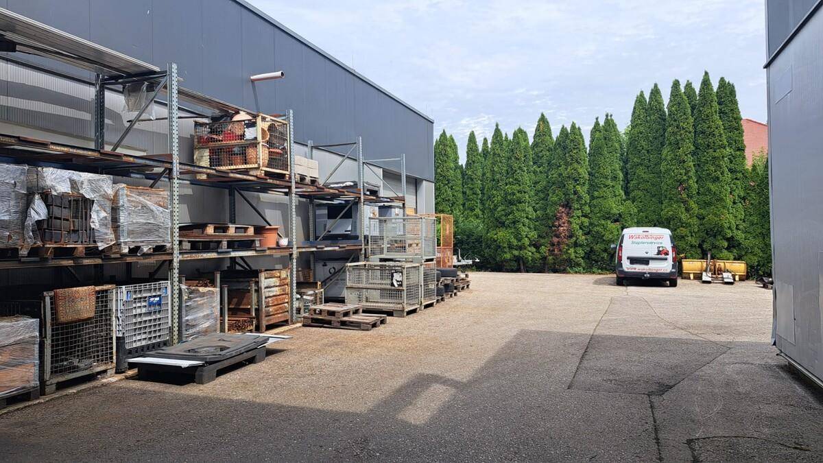 Weiterer Blick auf den Außenlagerbereich mit Regalen voller Waren, einem Lieferwagen und hohen Bäumen.