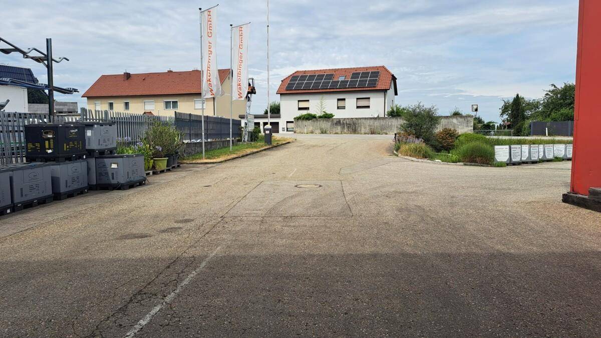 Gepflasterter Außenbereich, möglicherweise ein Parkplatz, mit Gebäuden im Hintergrund, eines mit Solaranlage.