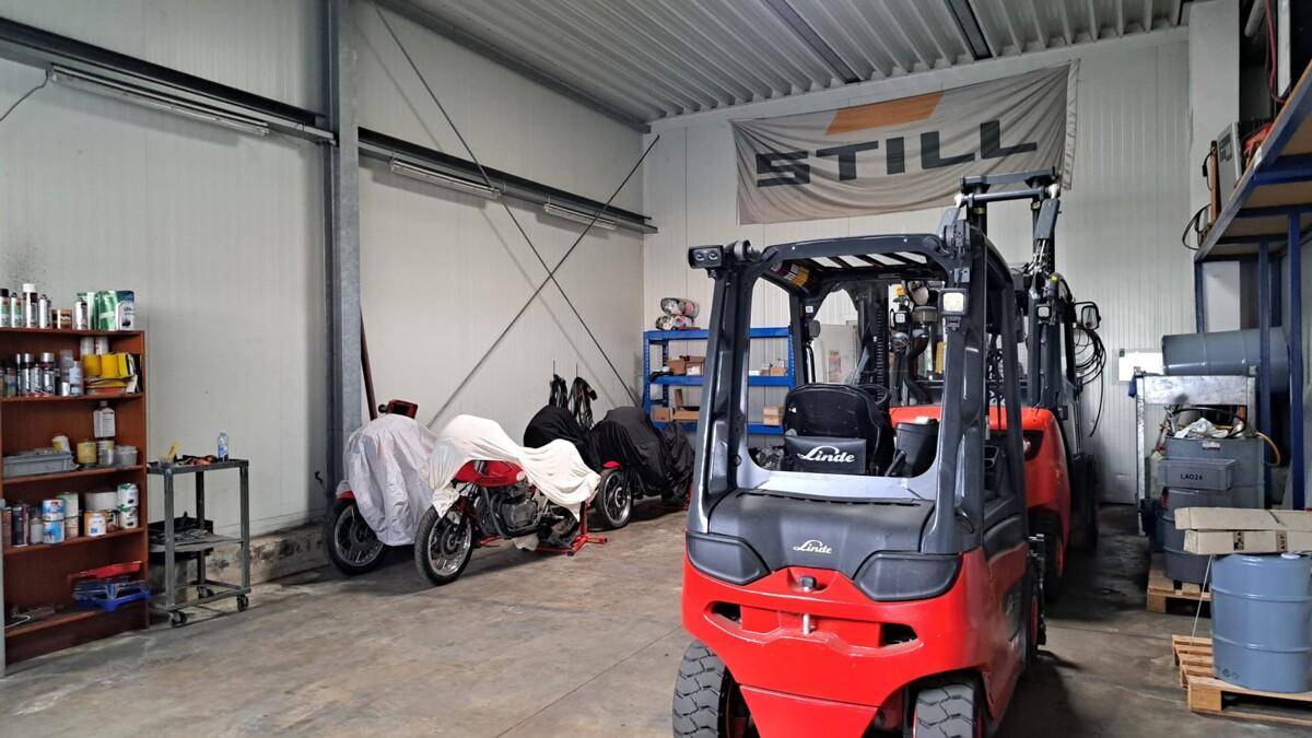 Innenansicht einer Lagerhalle oder Werkstatt mit Gabelstapler, abgedeckten Motorrädern und Regalsystemen.