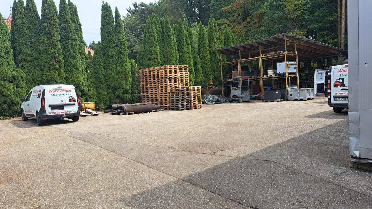 Außenlagerbereich mit gestapelten Paletten, Lieferwagen und einer Reihe hoher Nadelbäume im Hintergrund.