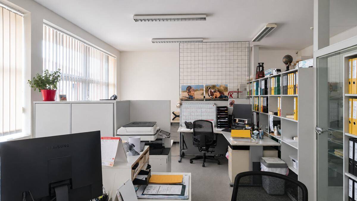 Großraumbüro mit mehreren Schreibtischen, Bürostühlen, Regalen voller Akten und Fenstern mit Jalousien.