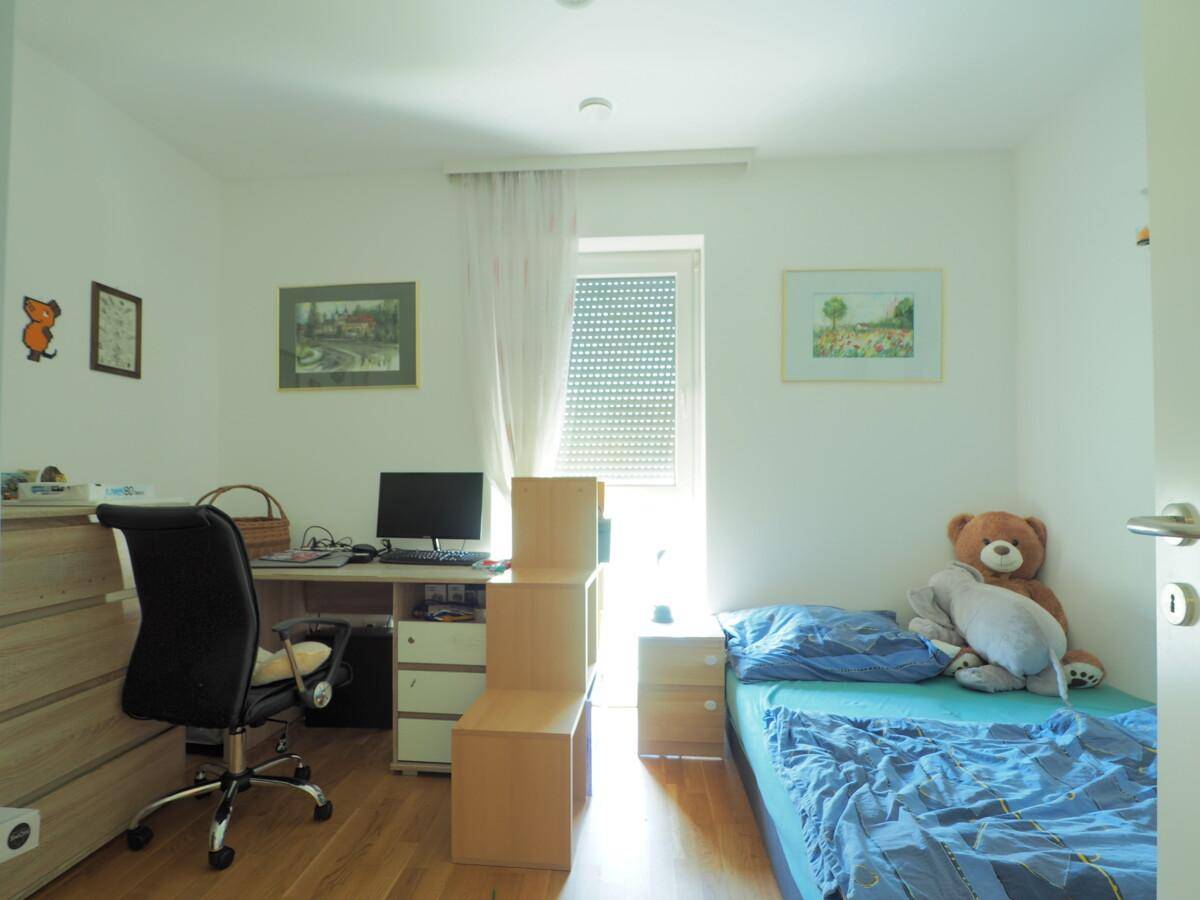 Kinderzimmer mit Einzelbett, Schreibtisch, Holzboden und Fenster mit Rollladen.