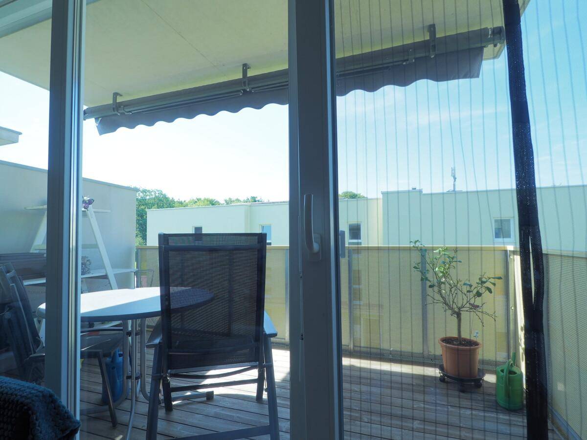 Balkon mit Holzboden, Markise und Sitzgelegenheiten, umgeben von anderen Gebäuden.