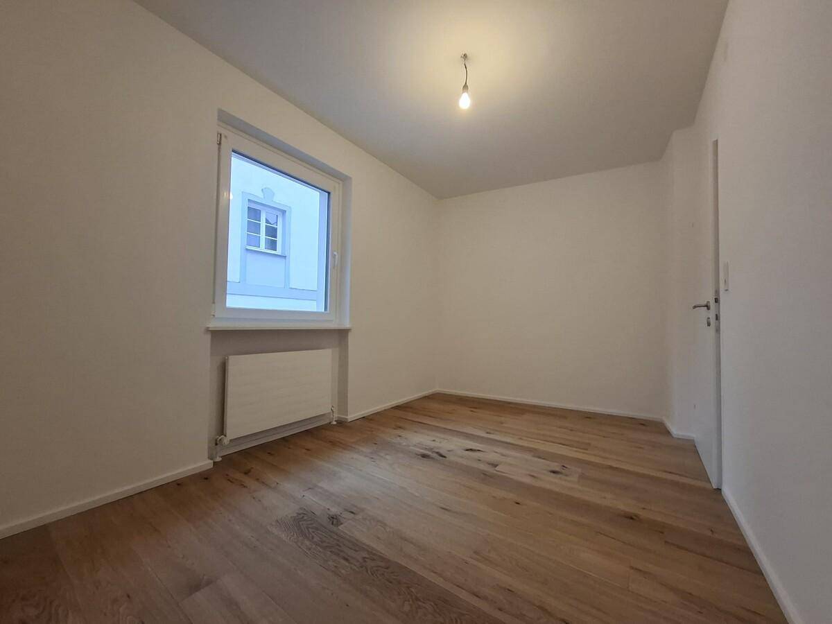 Helles Schlafzimmer mit Holzboden und Fenster, das auf ein gegenüberliegendes Gebäude blickt.