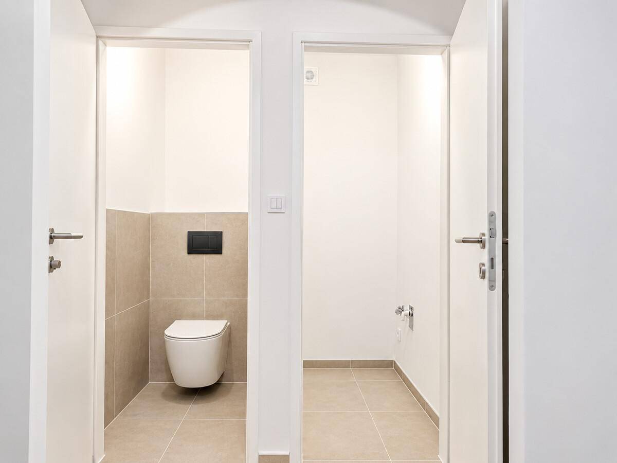 Separates WC mit Wand-WC und beigefarbenen Fliesen, daneben ein weiterer Raum.
