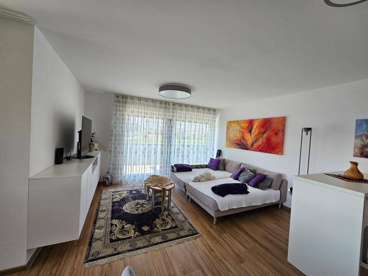 Helles Wohnzimmer mit beigem Sofa, Teppich und großen Fenstern mit Blick ins Grüne.
