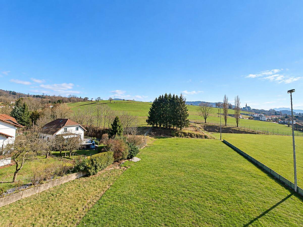 Weitläufige grüne Landschaft mit Hügeln, Bäumen und einigen Häusern in der Ferne.
