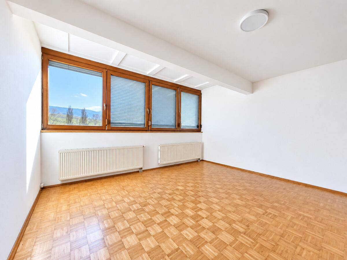 Helles Zimmer mit Parkettboden, großen Holzfenstern und Heizkörpern, ideal als Wohn- oder Schlafzimmer.
