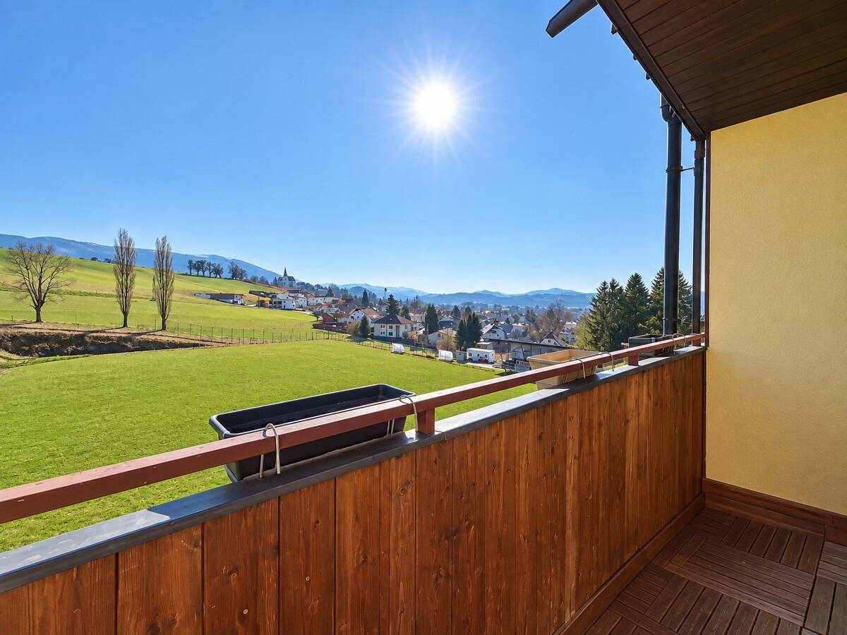 Sonniger Balkon mit Holzboden und weitem Blick über grüne Felder und ein Dorf.