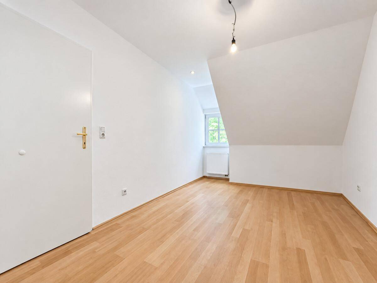 Leeres Zimmer im Dachgeschoss mit Parkettboden, schrägen Wänden und Fenster.