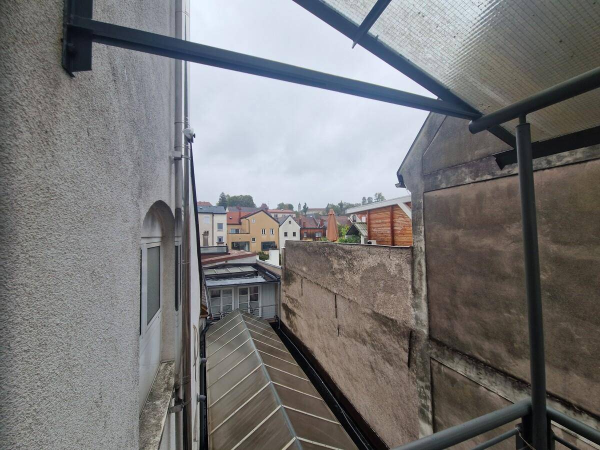 Blick von einem Balkon in eine enge Gasse zwischen Gebäuden, mit weiteren Dächern im Hintergrund.