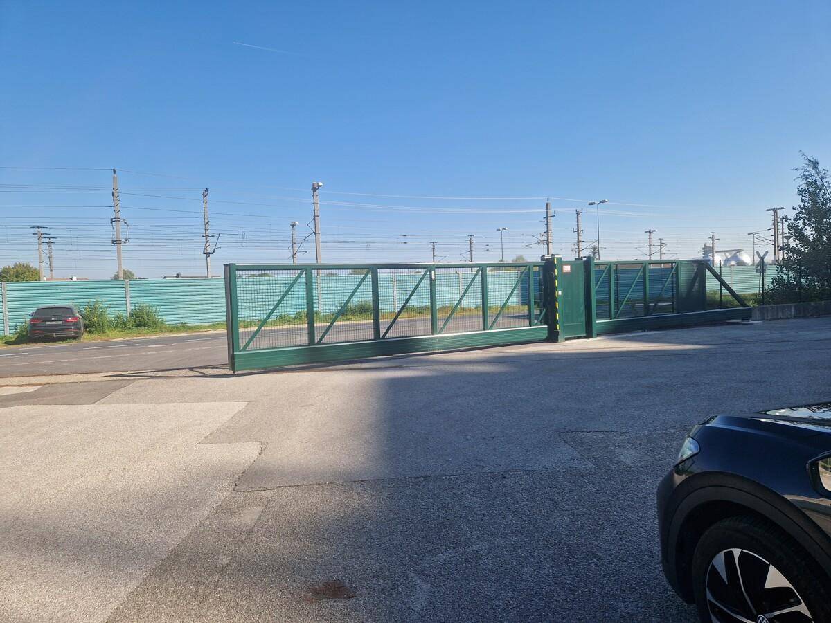 Großes, grünes Metallschiebetor, das den Zugang zu einem gepflasterten Bereich ermöglicht, mit Stromleitungen im Hintergrund.