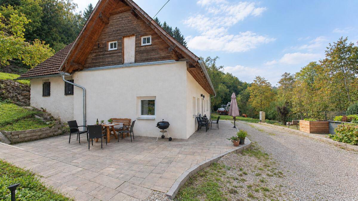 Hausfassade mit großer Terrasse, Sitzmöbeln und Grill, eingebettet in eine grüne Hügellandschaft.