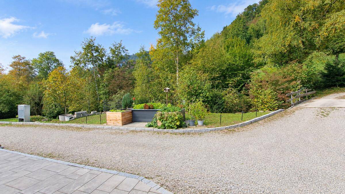Kiesauffahrt mit gepflegtem Gartenbereich und Blick auf die bewaldeten Hügel.