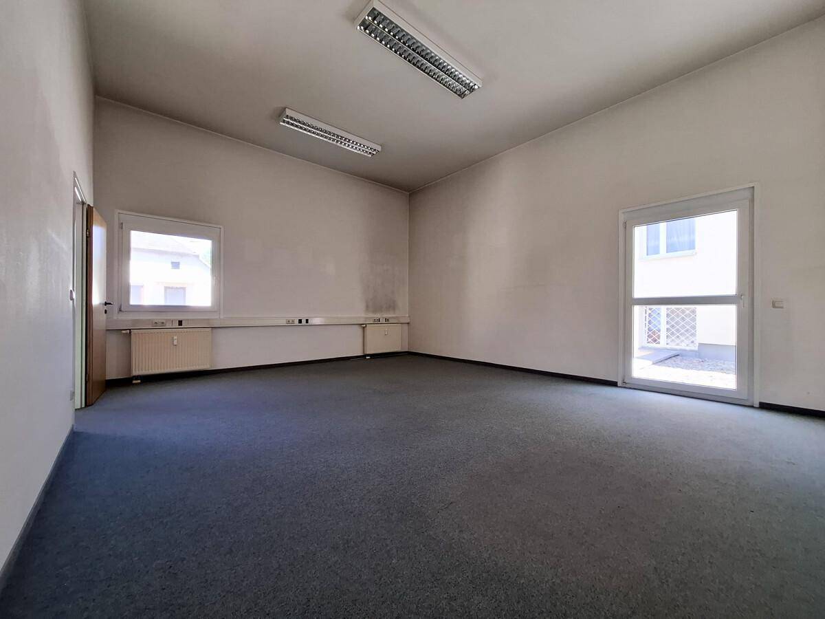 Helles, leeres Büro mit Teppichboden und Zugang zu einem Außenbereich durch eine Glastür.