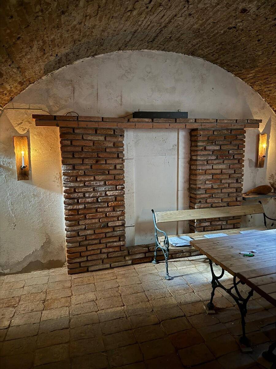Gewölbekeller mit Ziegelwandelementen, Holztisch und Bank, beleuchtet von Wandleuchten.