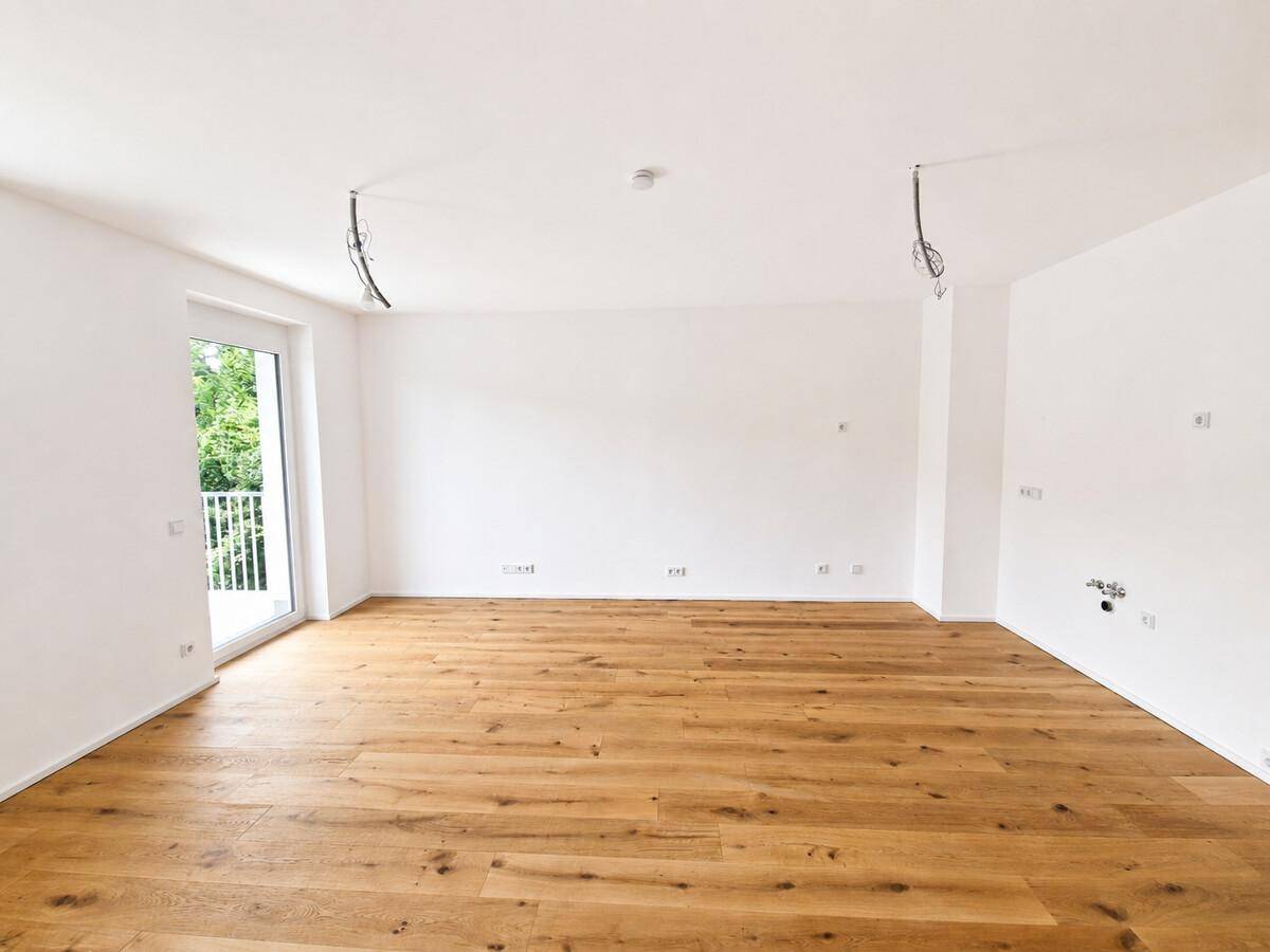 Helles, leeres Wohnzimmer mit Holzboden und Zugang zu einem Balkon.