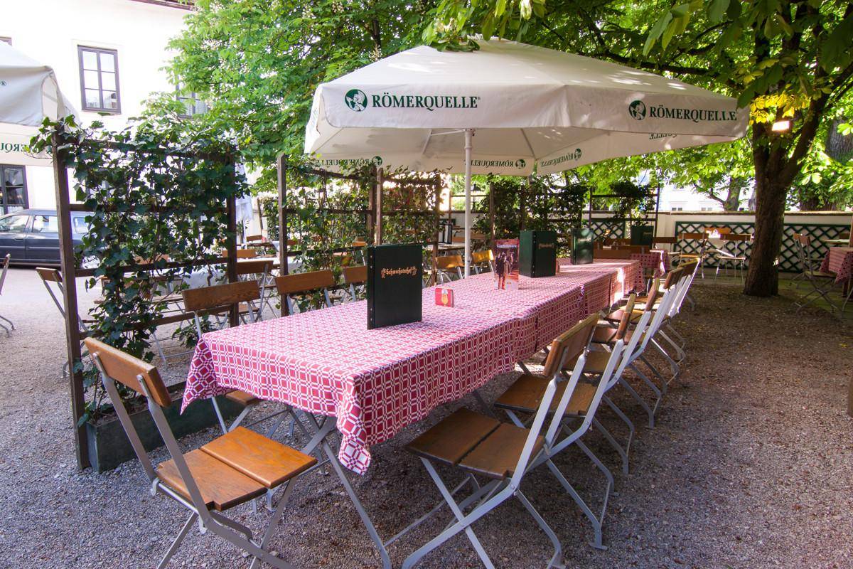 Schattiger Biergarten mit langen Tischen, Stühlen und großem Sonnenschirm unter Bäumen.