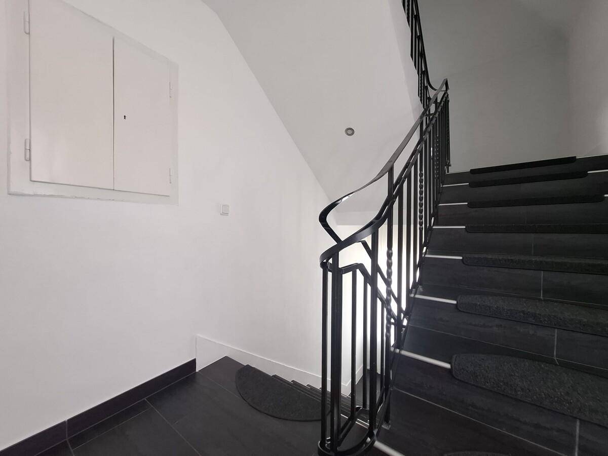 Treppenhaus mit dunklen Stufen, schmiedeeisernem Geländer und weißen Wänden.