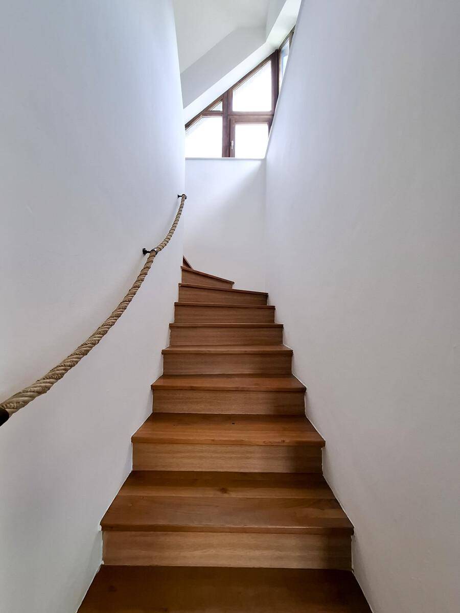 Holztreppe mit Seilgeländer führt nach oben zu einem hellen Fensterbereich.