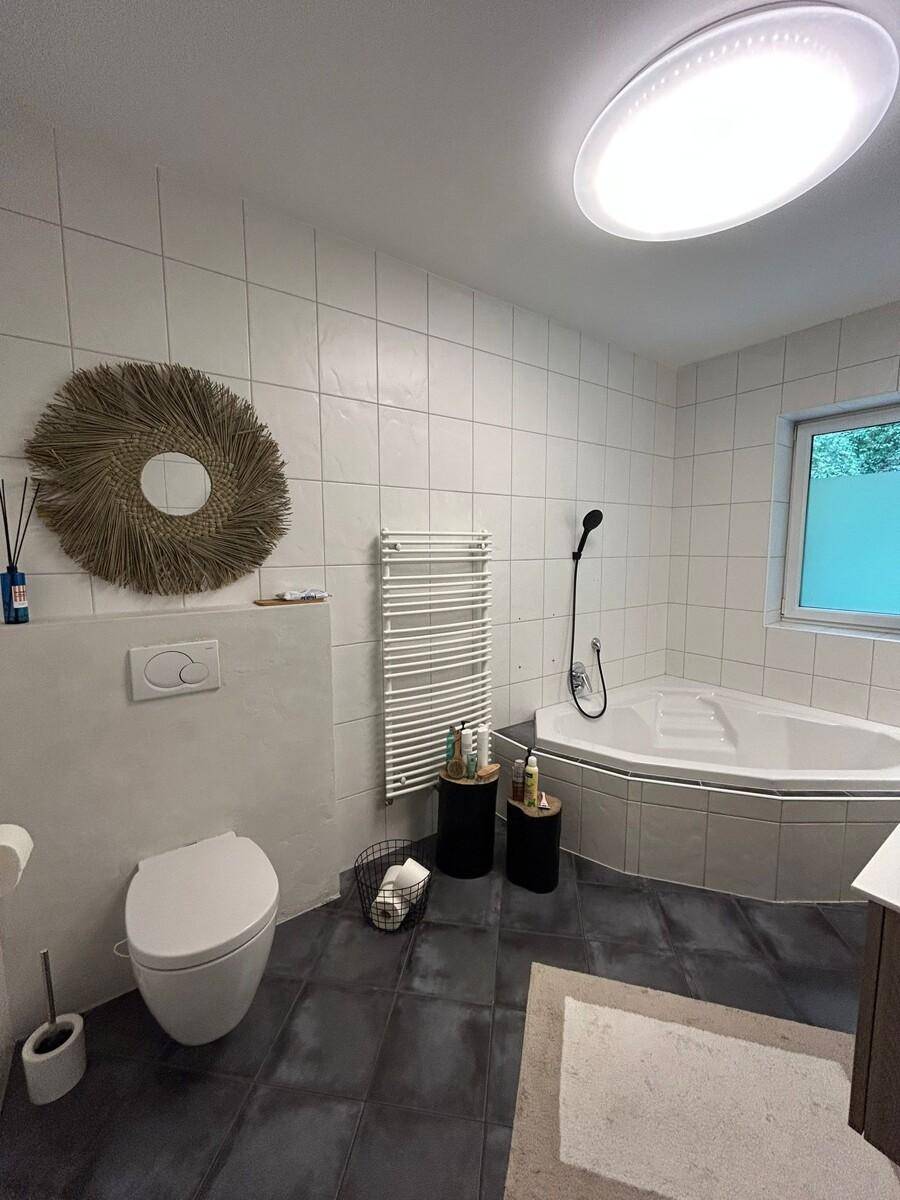 Modernes Badezimmer mit Eckbadewanne, Wand-WC und Handtuchheizkörper, hell gefliest.