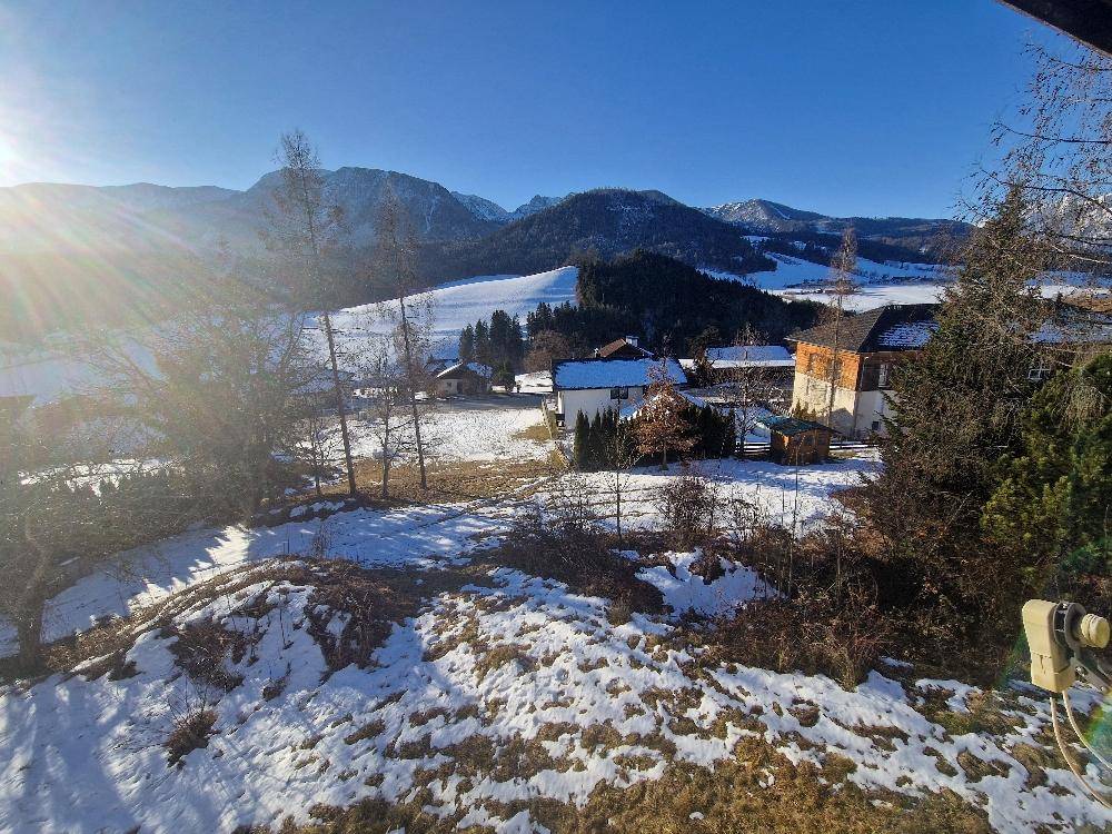 Weitläufige Winterlandschaft mit schneebedeckten Feldern, Häusern und Bergen unter klarem blauem Himmel.