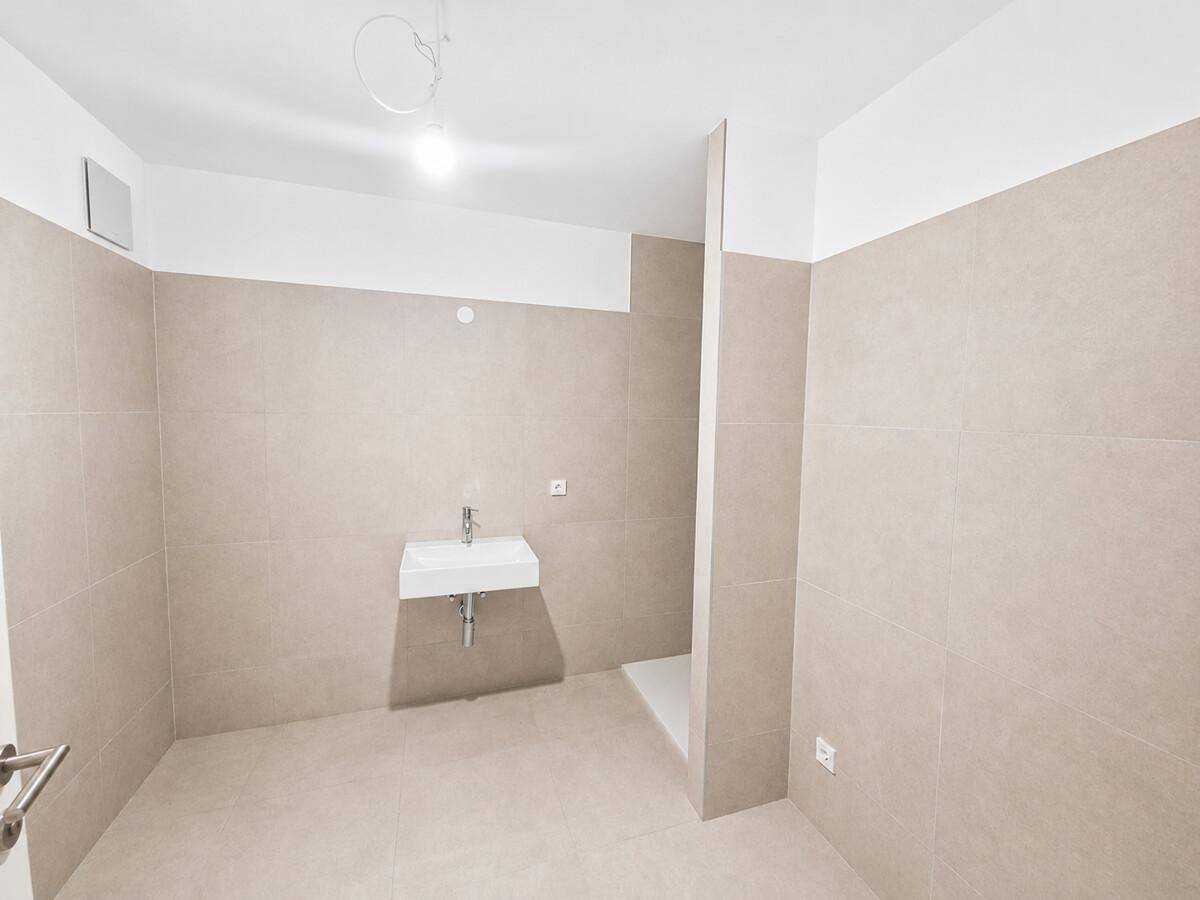 Unmöbliertes Badezimmer mit Wandwaschbecken, Duschbereich und beigefarbenen Fliesen.