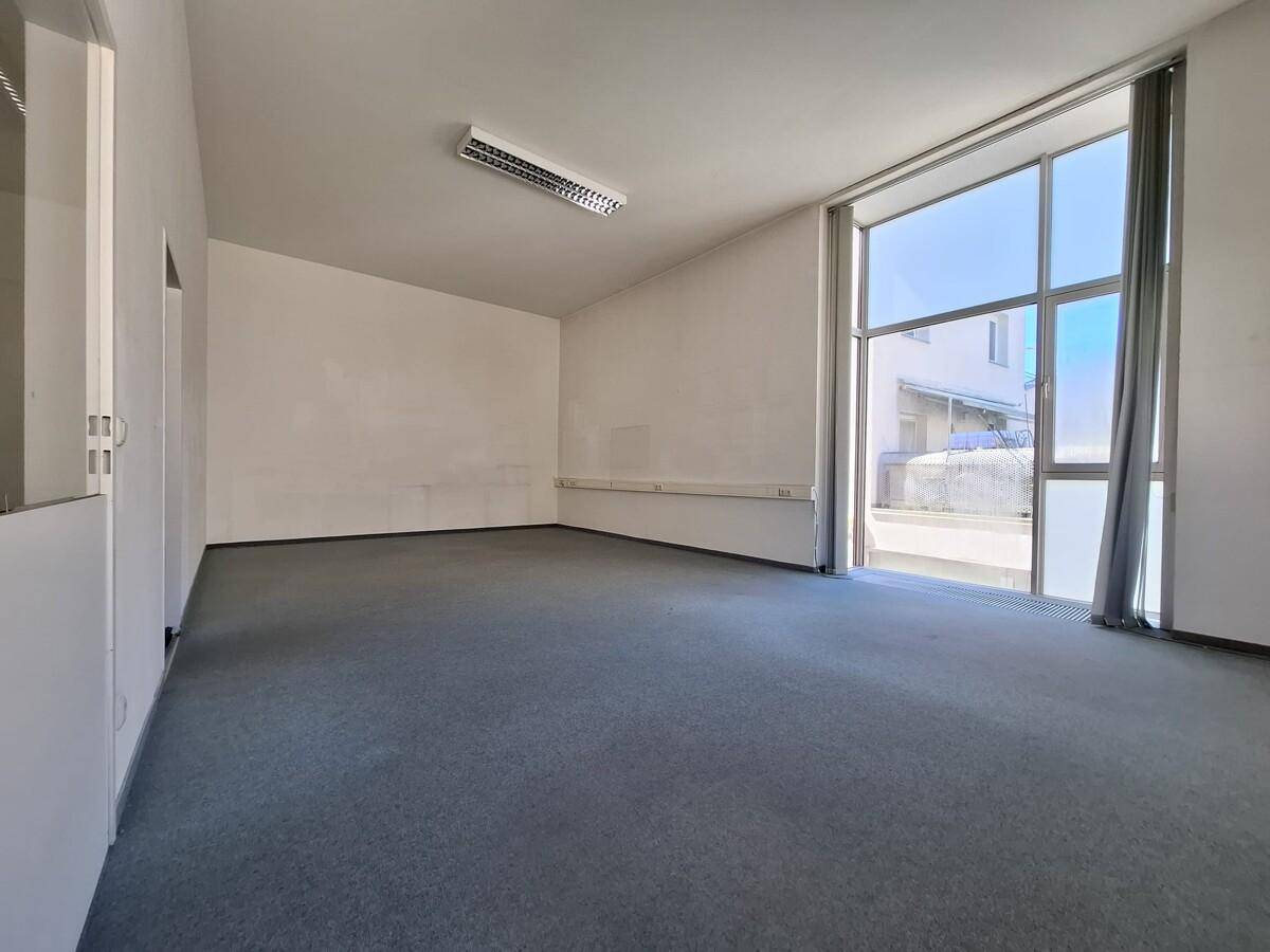 Leeres Büro mit Teppichboden und einer großen Fensterfront, die den Raum hell erscheinen lässt.