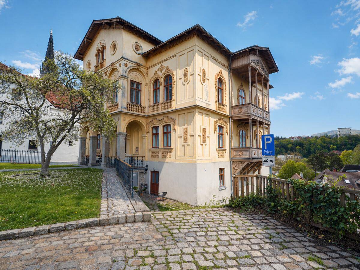 Seitenansicht des historischen Gebäudes mit dekorativem Holzbalkon und gepflastertem Zugang.