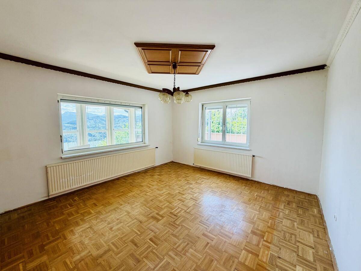 Helles Zimmer mit Parkettboden, zwei Fenstern und weitem Blick auf die grüne Hügellandschaft.