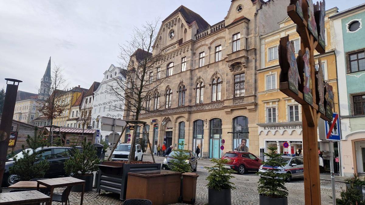 Belebte Straße mit historischen Gebäuden, geparkten Autos und Außengastronomie in einer Altstadt.
