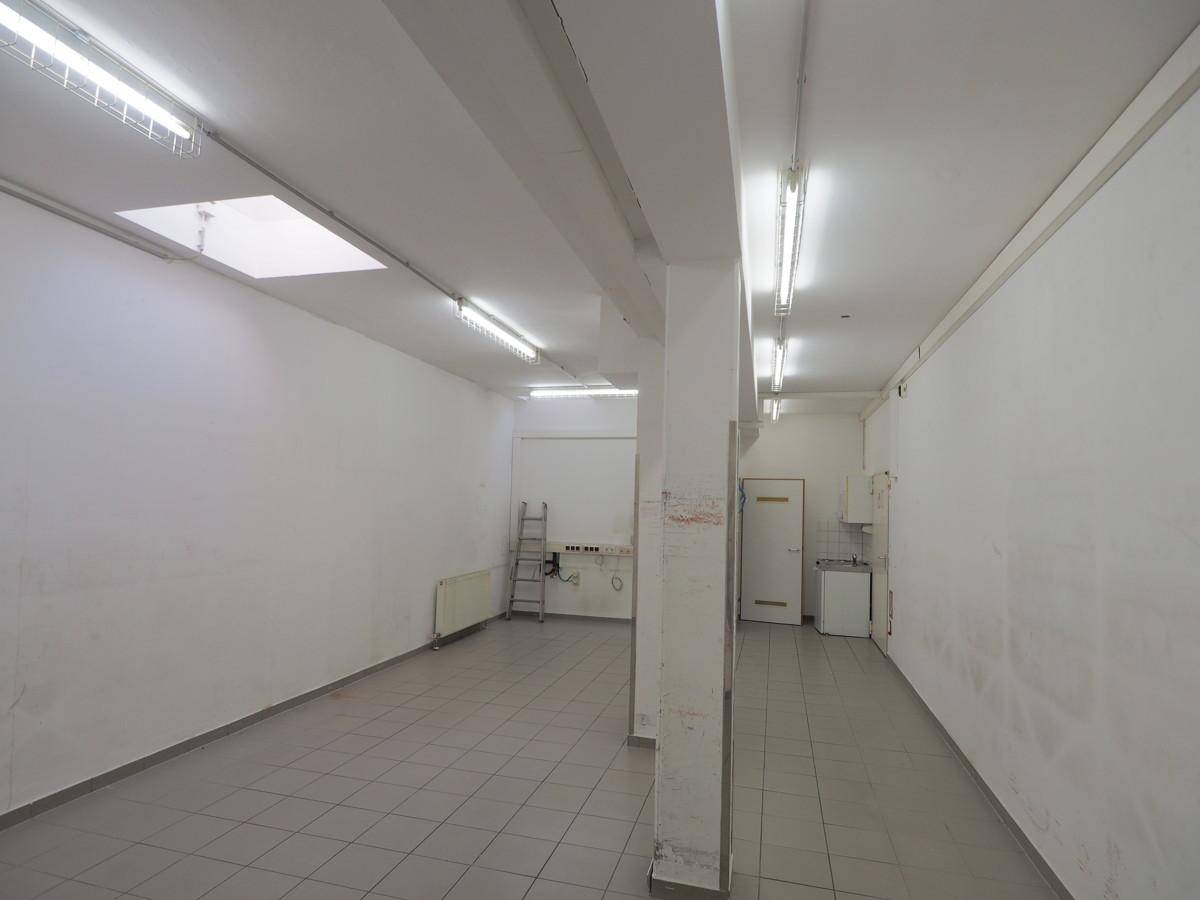 Leerer Gewerberaum mit Fliesenboden, weißen Wänden, Leuchtstoffröhren und einem kleinen Waschbeckenbereich.