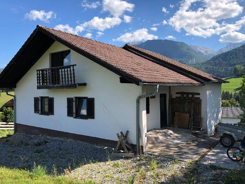 Ländliches Haus mit weißer Fassade, braunem Dach und kleinem Holzbalkon vor einer Bergkulisse.