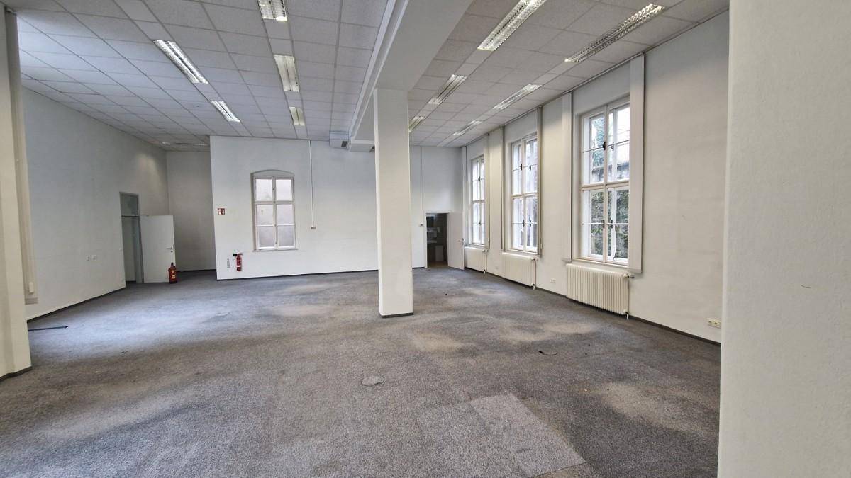 Großer, offener Büroraum mit Teppichboden, Säulen und mehreren hohen Fenstern, die den Raum erhellen.