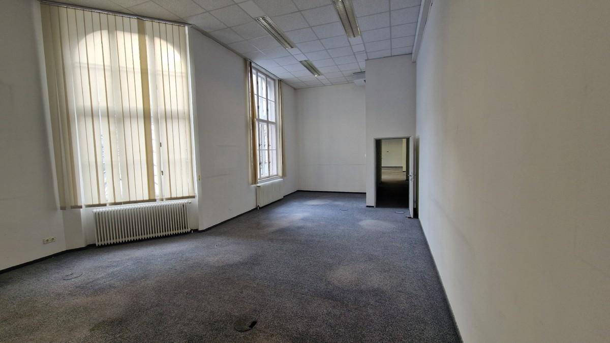 Leerer Büroraum mit Teppichboden und einem großen Fenster, das den Raum mit Tageslicht füllt.