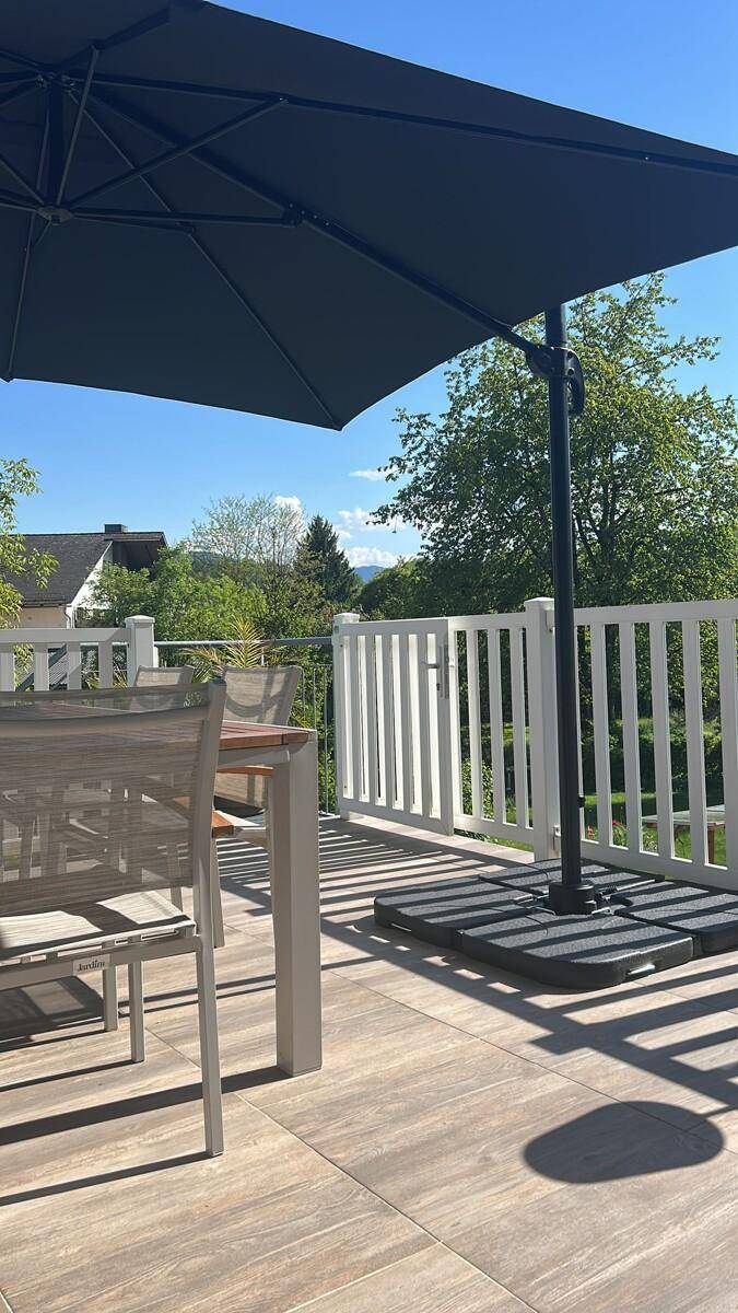 Sonniger Terrassenbereich mit Holzboden, Esstisch und Sonnenschirm, umgeben von einem weißen Geländer.