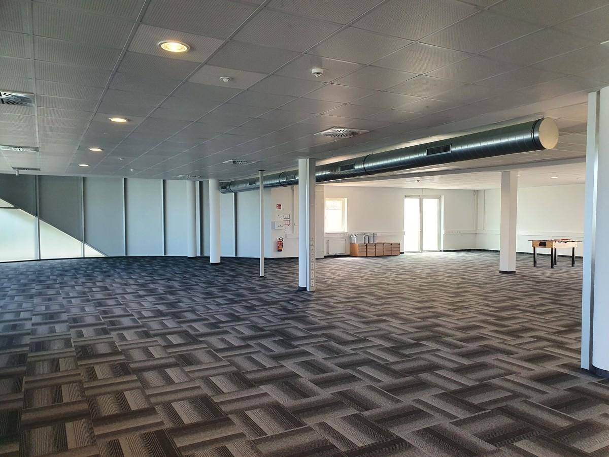 Geräumiger, offener Büroraum mit modularem Teppichboden, Säulen und sichtbaren Lüftungsleitungen an der Decke.
