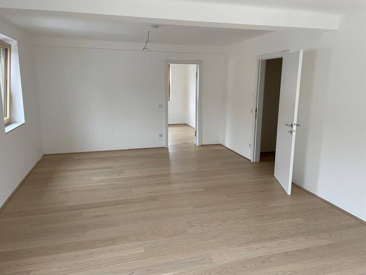 Geräumiges, leeres Wohnzimmer mit hellem Parkettboden und großen Fenstern.