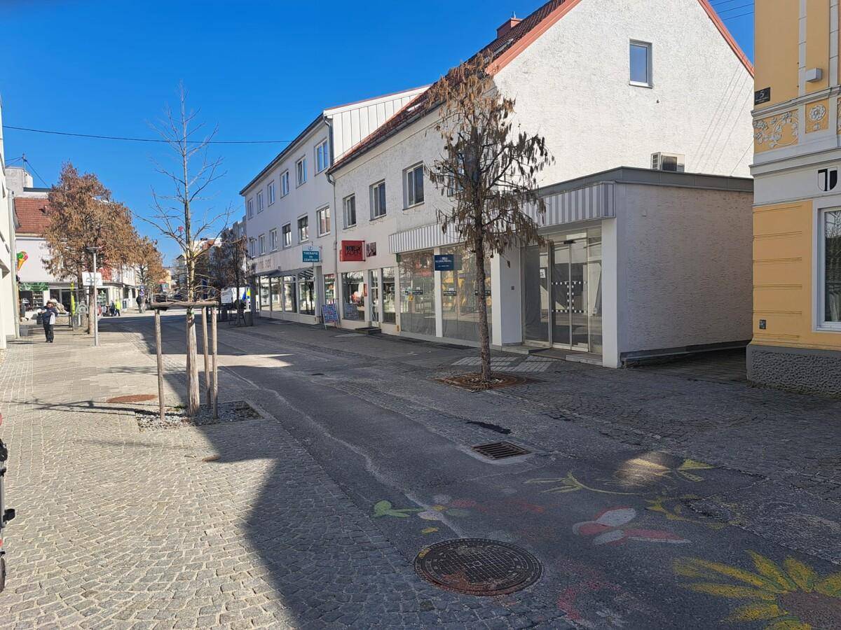 Seitenansicht einer Geschäftsstraße mit Gebäuden, Gehwegen und Kreidezeichnungen auf dem Bürgersteig unter blauem Himmel.