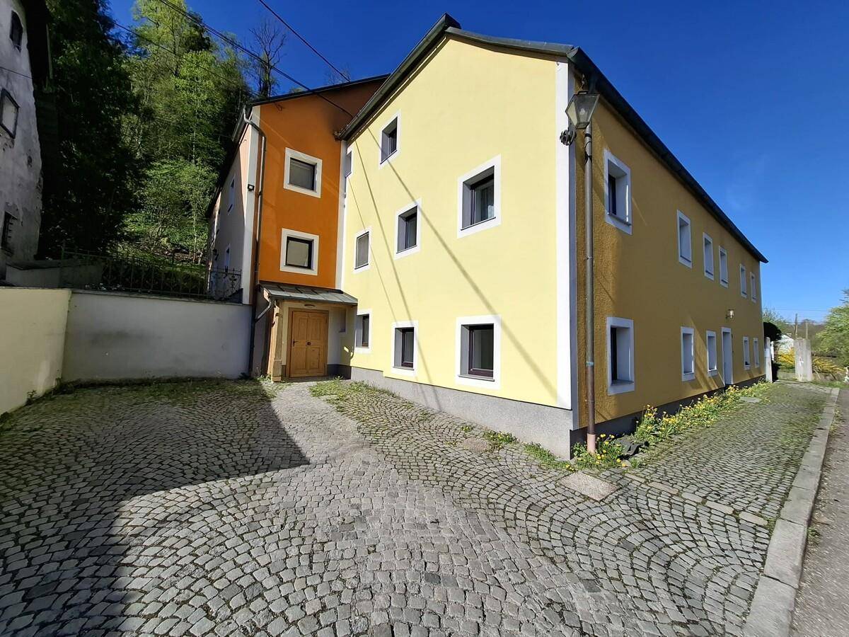 Gelbes Gebäude mit gepflastertem Vorplatz und grüner Umgebung unter blauem Himmel.